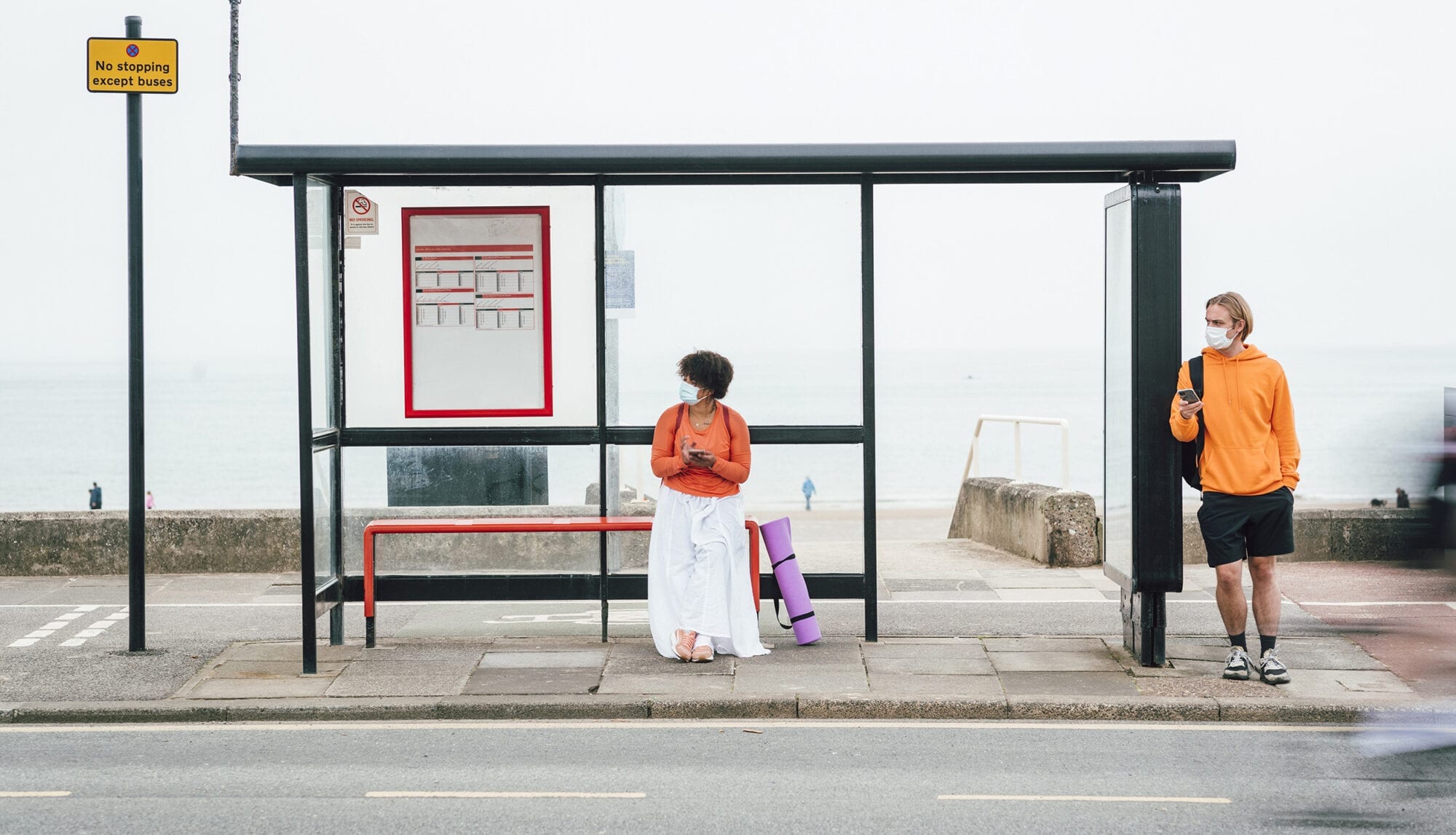 A woman of colour waits at a bus shelter by a road