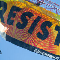 a huge orange and yellow banner filling the frame hanging from a crane reading RESIST with a person dangling off the left bottom corner