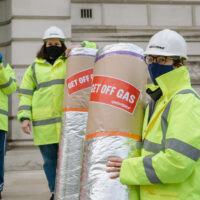 Two people in hard hats and reflective work jackets holding rolls of loft insulation, bearing labels reading 'Get off gas'