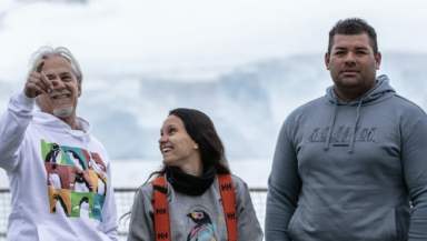 Three greenpeace crew members stand together on deck smiling and laughing. They're wearing Greenpeace sweaters and hoodies with various penguin designs