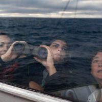 Three crew members look through binoculars out of a window. The ocean and evening sky are reflected in the window.