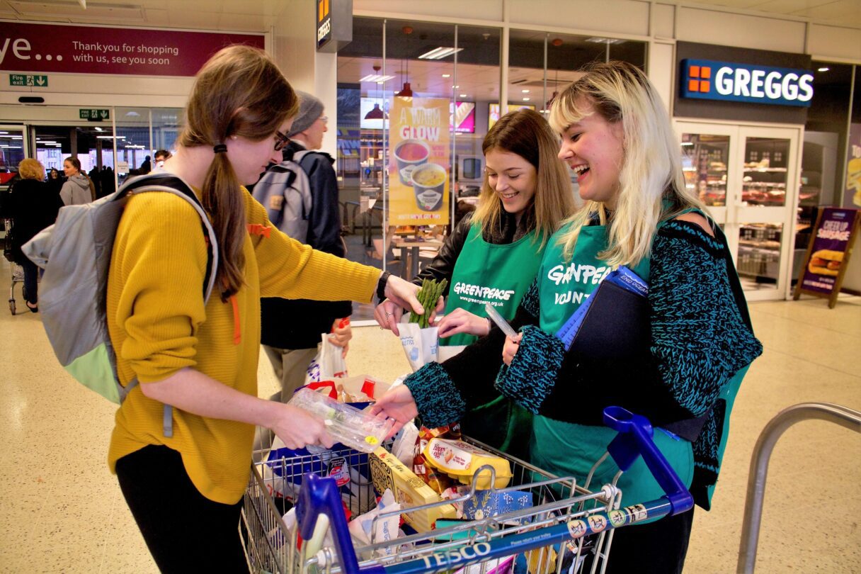 Two smiling Greenpeace volunteers speak to a member of the public in a supermarket.