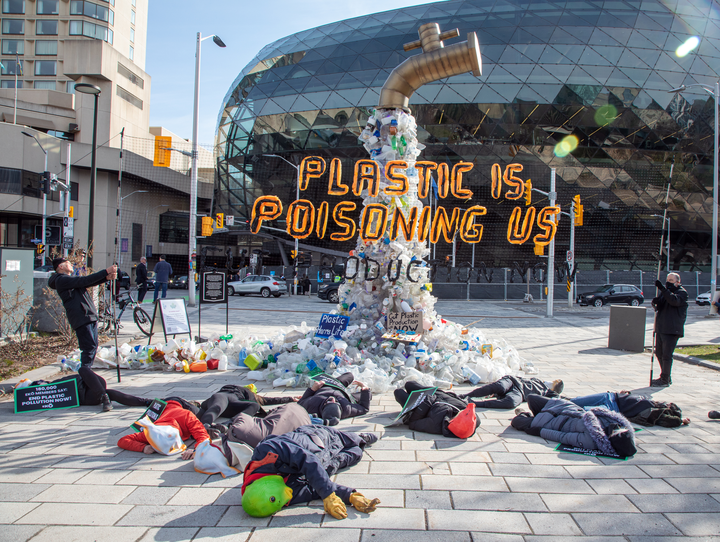 Giant sculpture of a tap with plastic pouring out. Banner in front says 'plastic is poisoning us'. protestors lie down in the foreground.