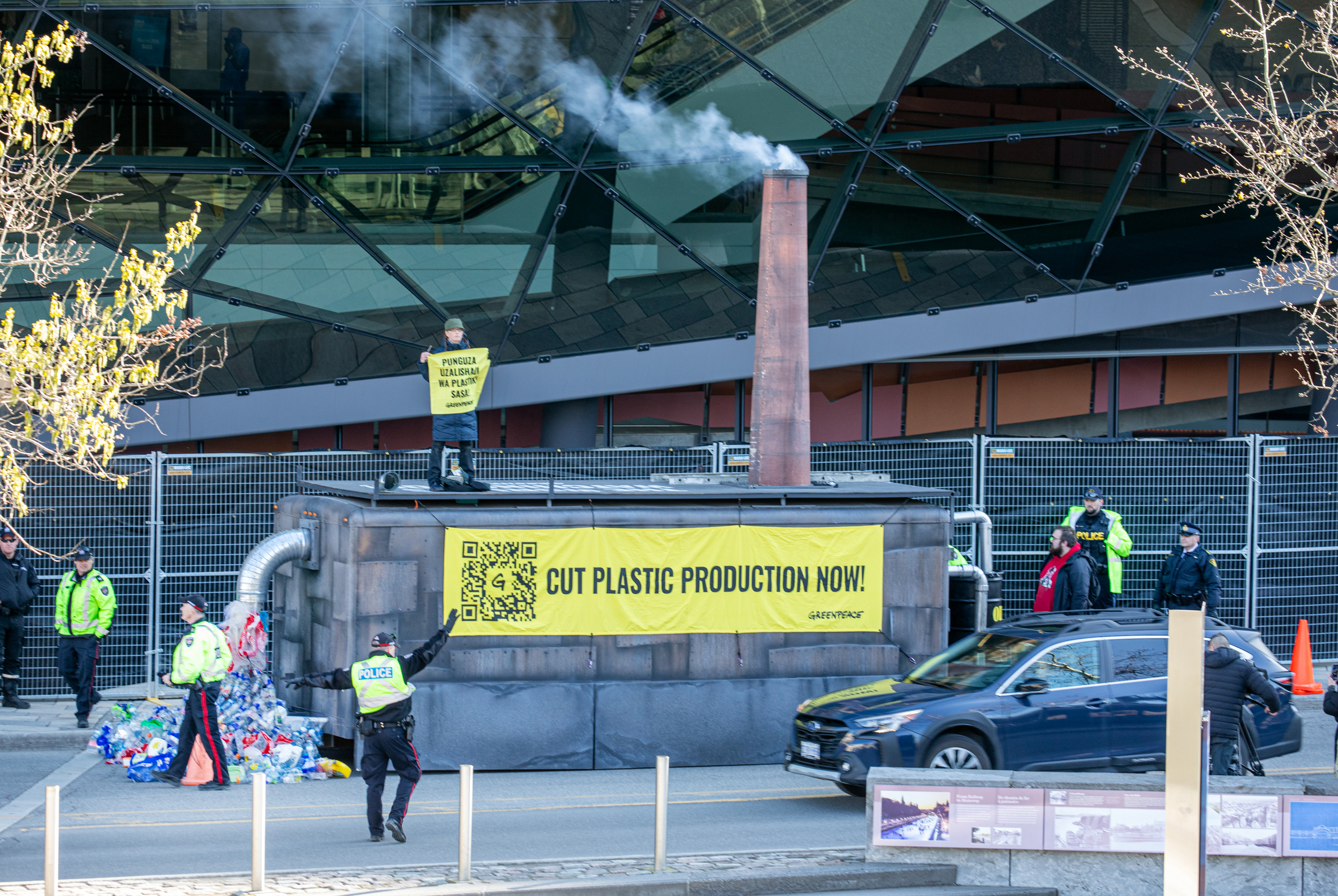 Mock factory with a smoking smokestack with a banner saying 'cut plastic production now'. Police in the foreground look bemused.