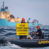 Two Greenpeace activists in an inflatable boat on the ocean, with a big ship in the background. One stands holding a a banner that says "Stop deep sea mining", while the other drives the boat.