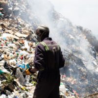 An African man in a boilersuit with hi-vis stripes and "care sanitation and suppliers ltd" written on the back of it stands looking out over a huge pile of smoking landfill rubbish towering over him. He is wearing a dirty face mask and a knitted cap, and thick gloves, holding crunched up Pepsi-branded plastic cups.
