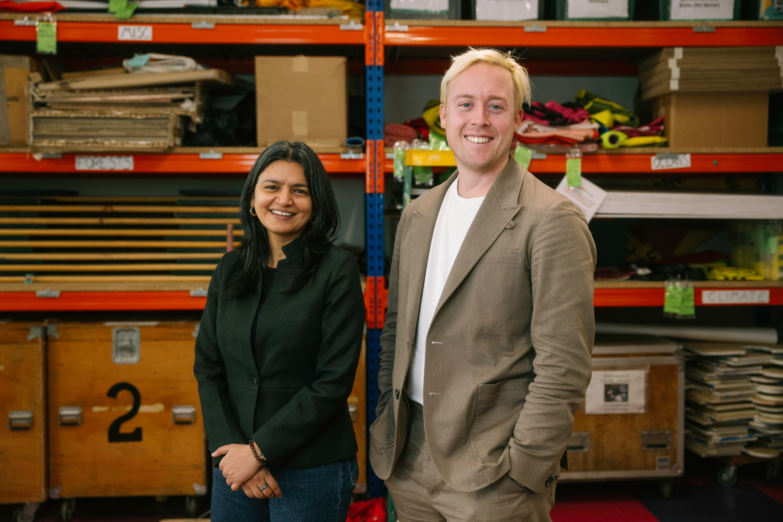 Relaxed portrait of Areeba Hamid and Will McCallum standing in front of a rack of shelves piled with campaigning paraphernalia, smiling into the camera.