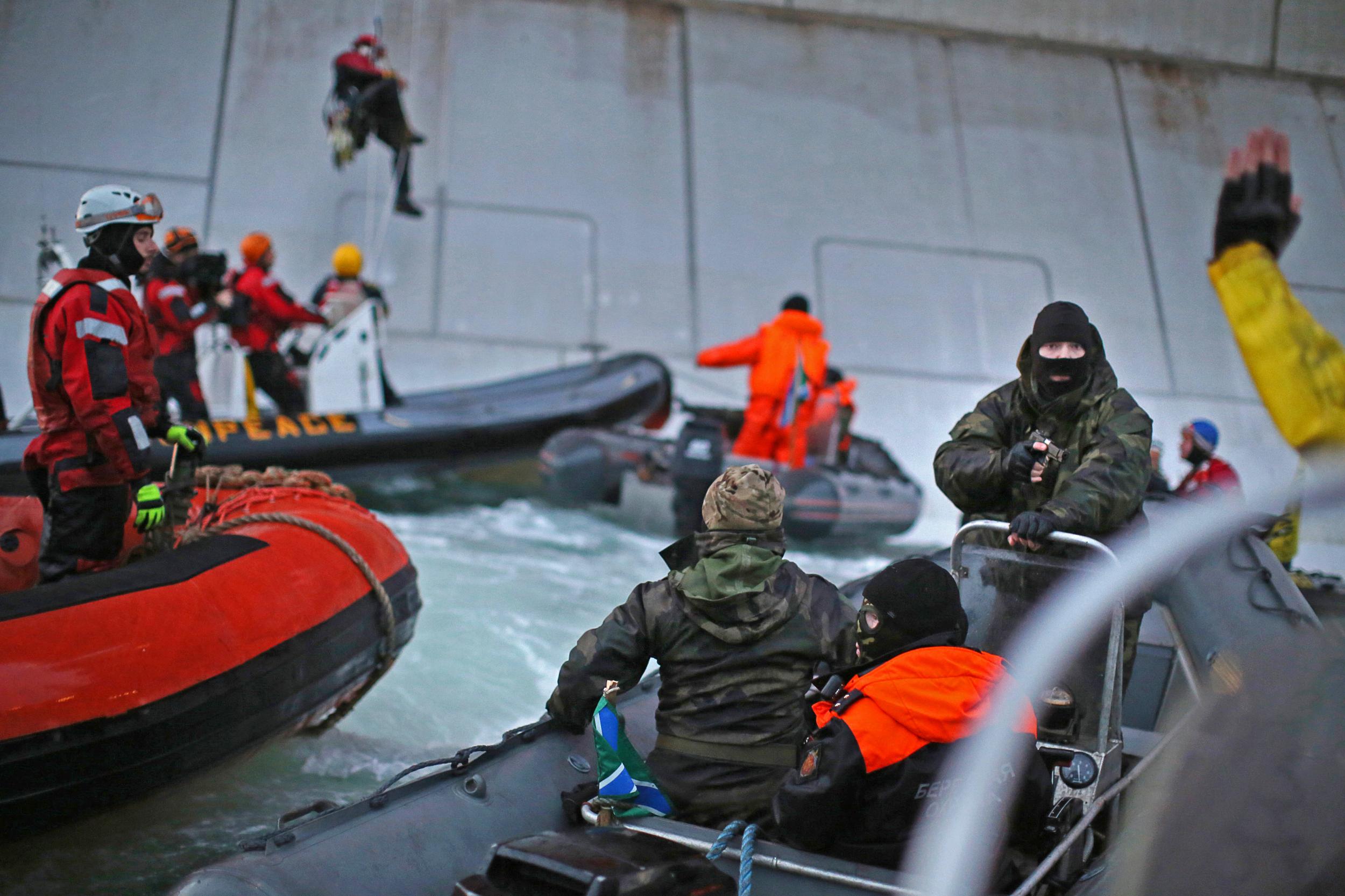 A man standing in a small boat in a black balaclava and camoflage jacket points a gun at someone just off-camera. Their hand can be seen raised in the air. In the background, more boats mill around, and a person is abseiling down the side of a large metal structure into a boat.