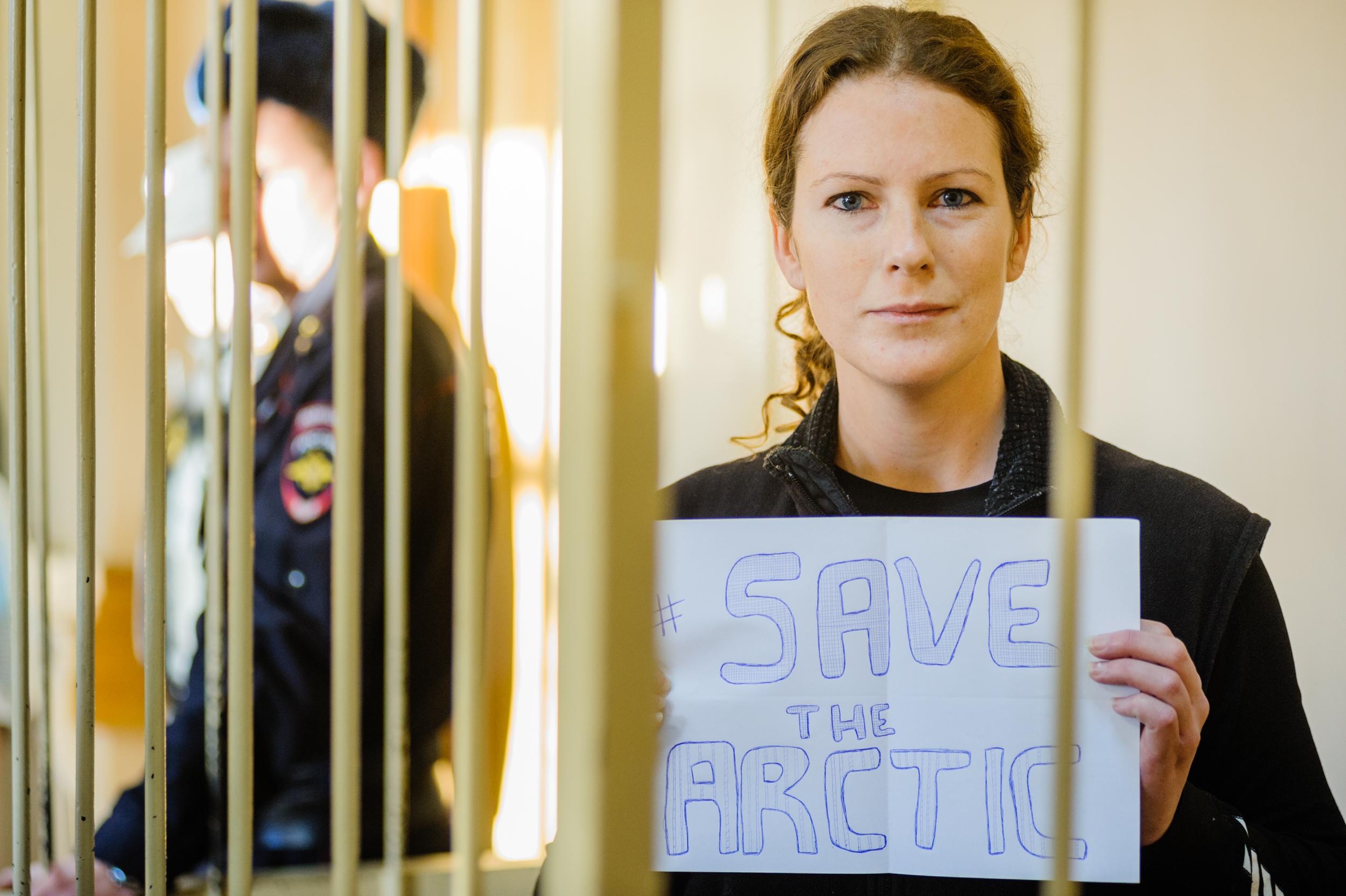 A woman behind bars looks into the camera with a serious expression. She holds up a sign saying 'save the Arctic'.