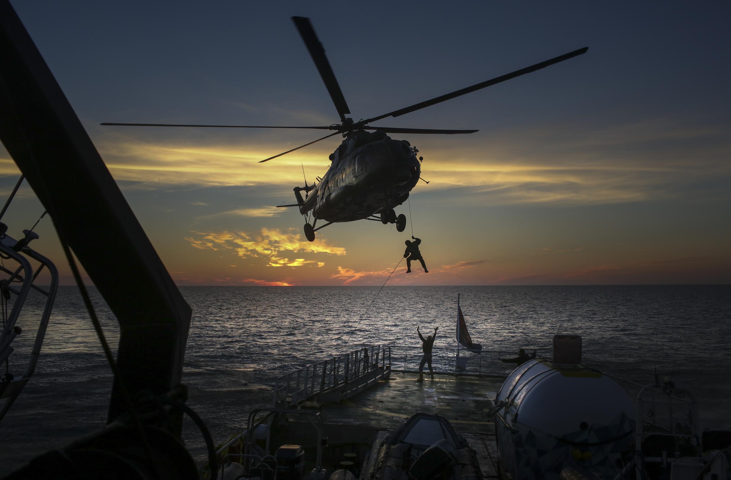 A helicopter is silhouetted against the dawn or dusk as it hovers low over the deck of a ship. A person is abseiling down onto the deck, where a crew member waves up at the helicopter with both hands.