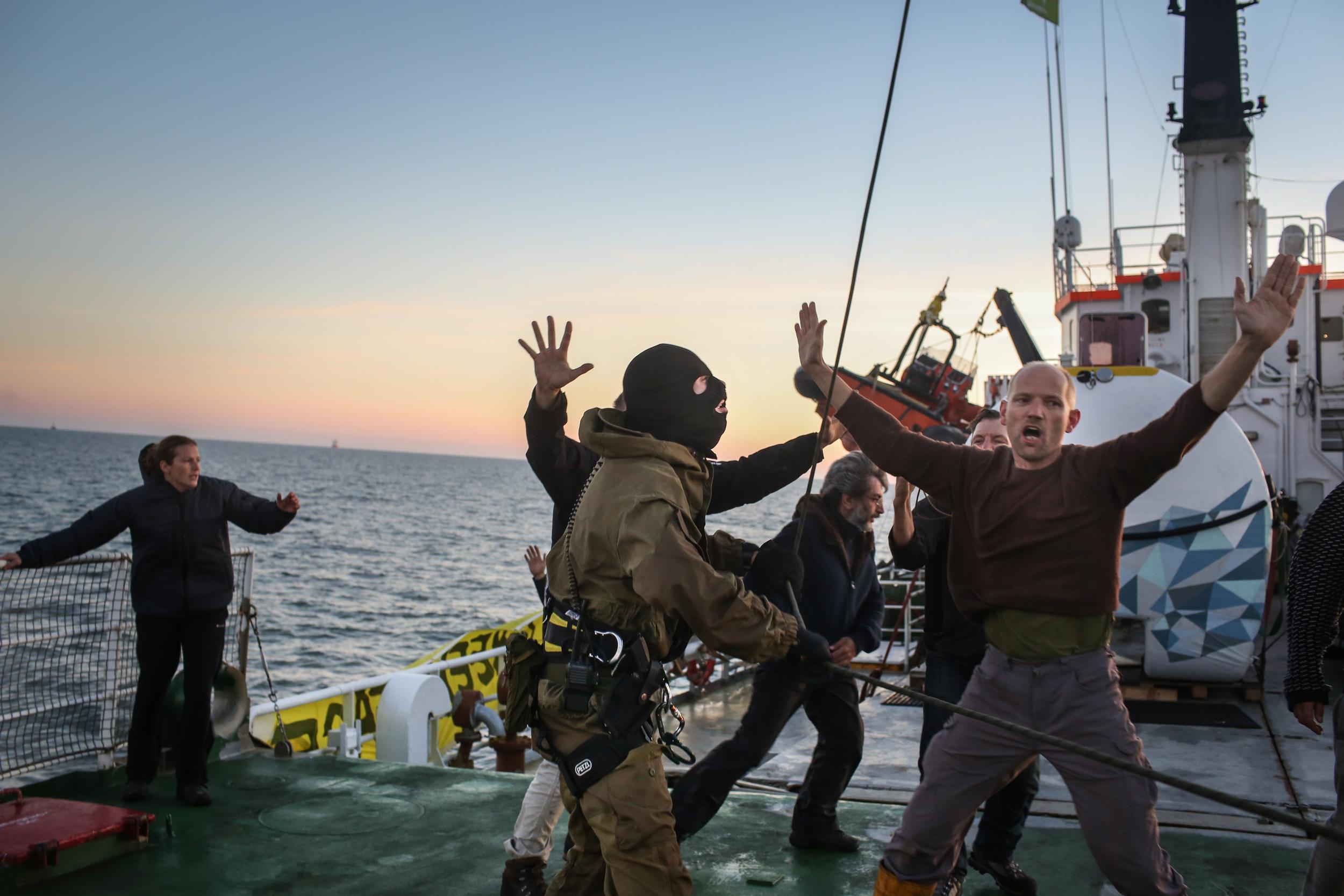 On the deck of a ship, a member of Greenpeace crew holds his hands up as he's accosted by a man in military uniform and a black balaclava.
