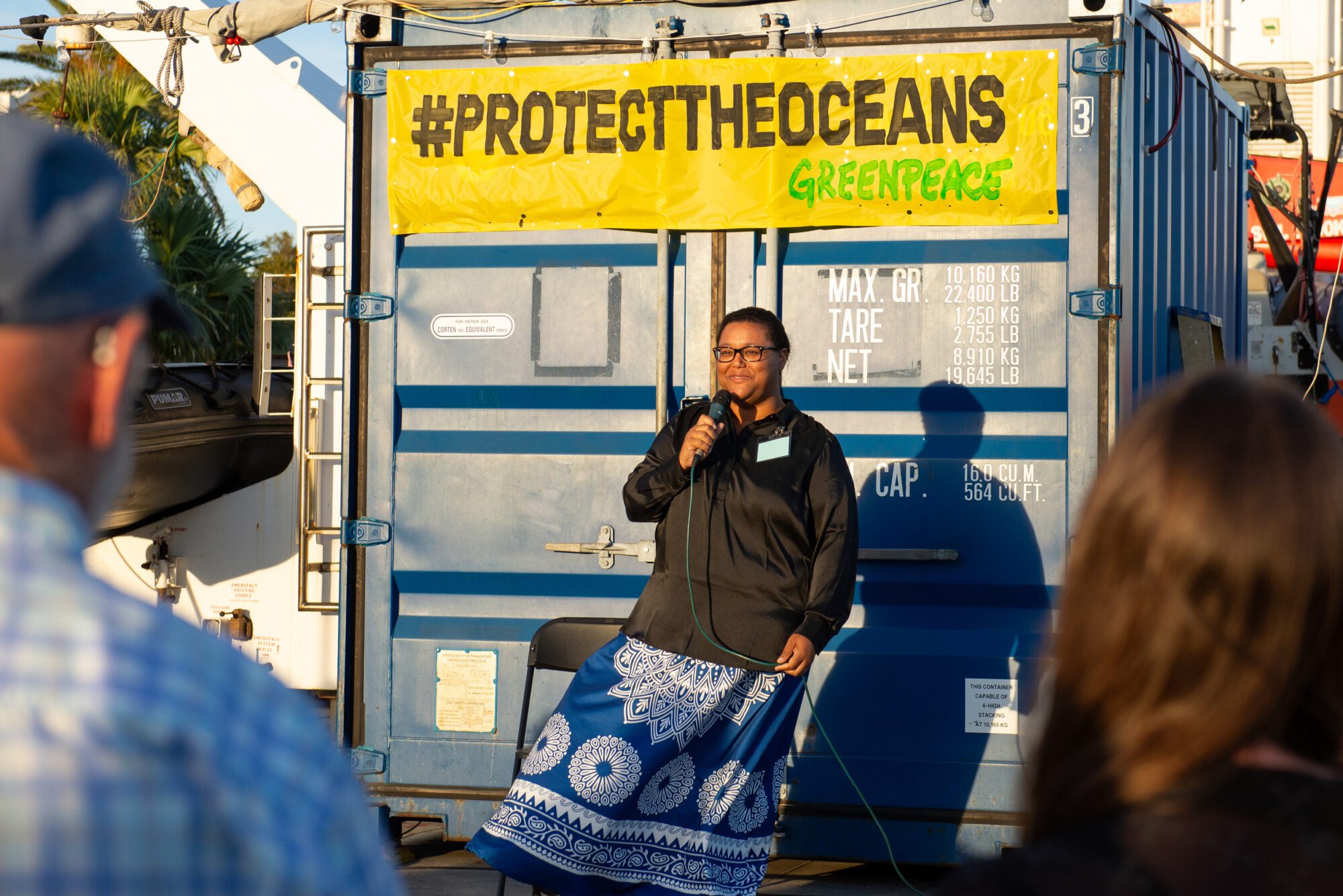 The author again holding a microphone in front of a blue metal container, with a yellow Greenpeace banner reading "Protect the Oceans" behind her, with a backs of a couple of audience members blurry in the foregroud