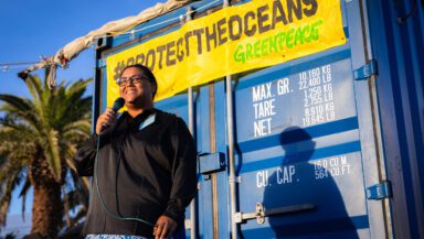 A woman of colour stands smiling and holding a microphone in front of a blue metal container, with a yellow Greenpeace banner reading "Protect the Oceans" behind her