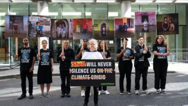 Activists in matching T-shirts with a Globe and "Climate Justice Now!" on them holding placards by Gideon Mendel of climate impacted communities from around the world and a large central banner at the front saying “Shell will never silence us on the climate crisis” protest outside the English Admirality Court in London.