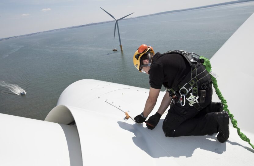 A worker in a hard hat and harness crouches on top of an offshore wind turbine. Other turbines are visible in the background.
