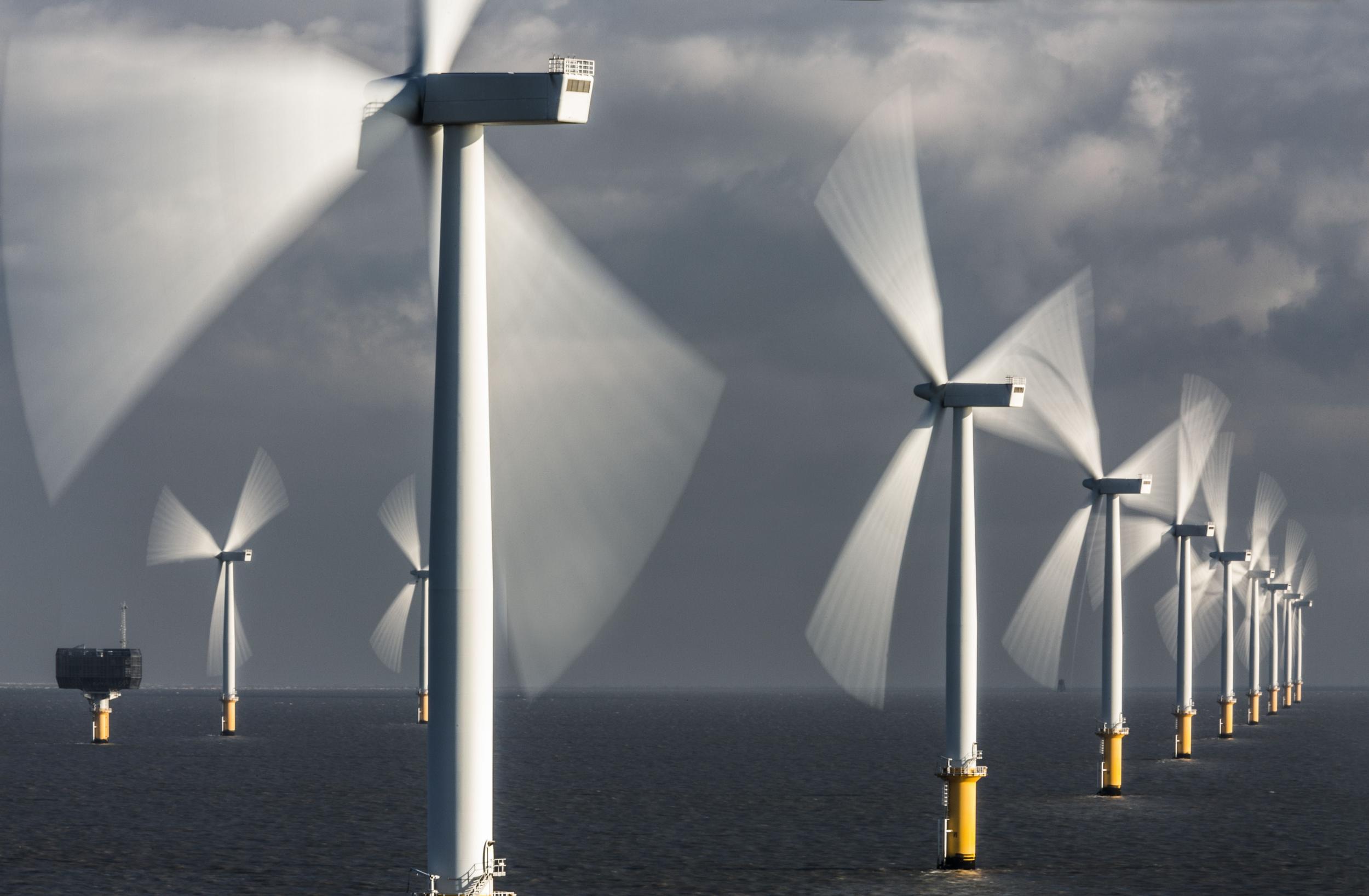 Offshore wind turbines lit by sun against a grey sky. Their blades are motion blurred.