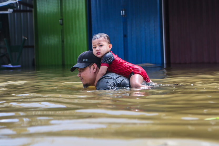 A toddler sitting on his father's shoulders as he wades through deep floodwater
