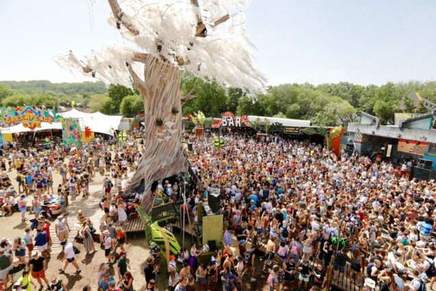A large tree with white wisps blows in the wind high above a festival field with many people in a crowd in it, and an colourful arch and signage for a bar