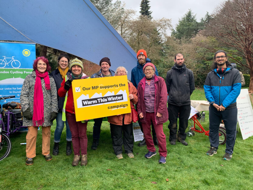 Nine people of mixed genders and races stand on grass in front of a tent with a sign next to it showing a bicycle icon. Two at the front hold a yellow sign reading 