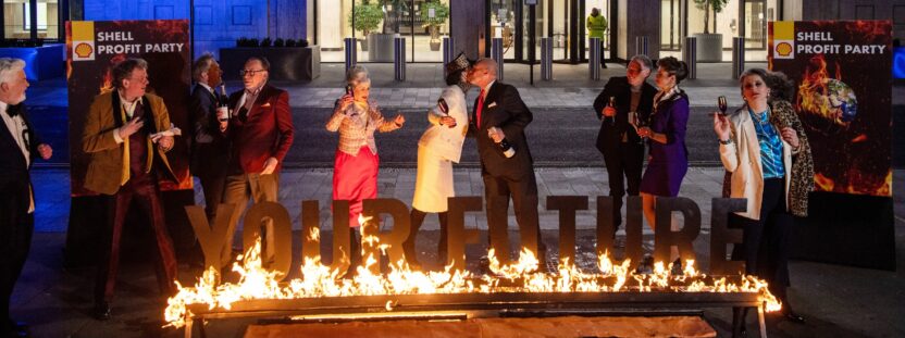 Activists dressed in suits perform as Shell shareholders. In the foreground, burning letters spell out 'our future'.