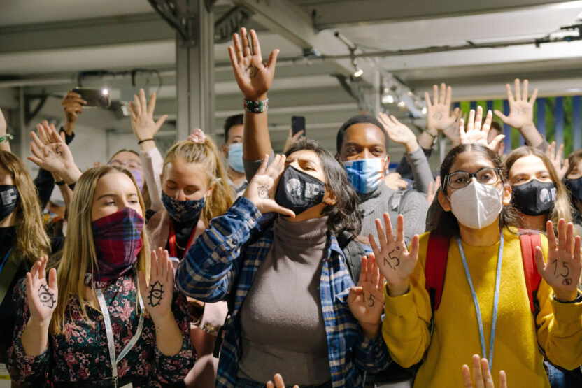 A group of masked young activists gathered round the camera. They're energetic and animated, with hands raised or cupped to their mouths as they chant slogans.