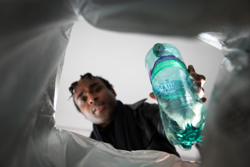 View from inside a bin, a person putting a green plastic bottle into it, framed by the plastic bag lining the bin.
