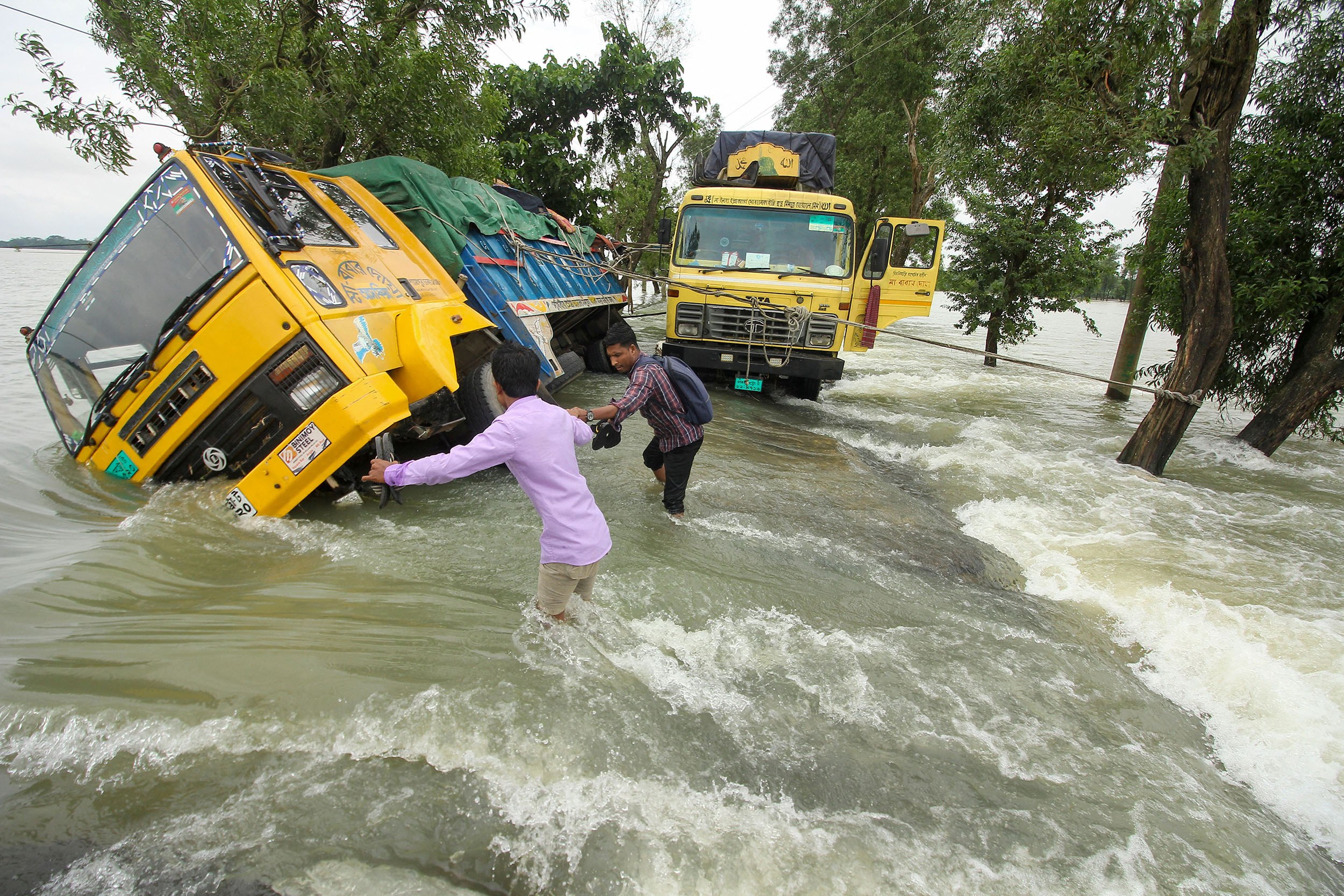 Two men wade through rushing water on a flooded road. They are passing two stranded trucks, one of the has fallen to its side.