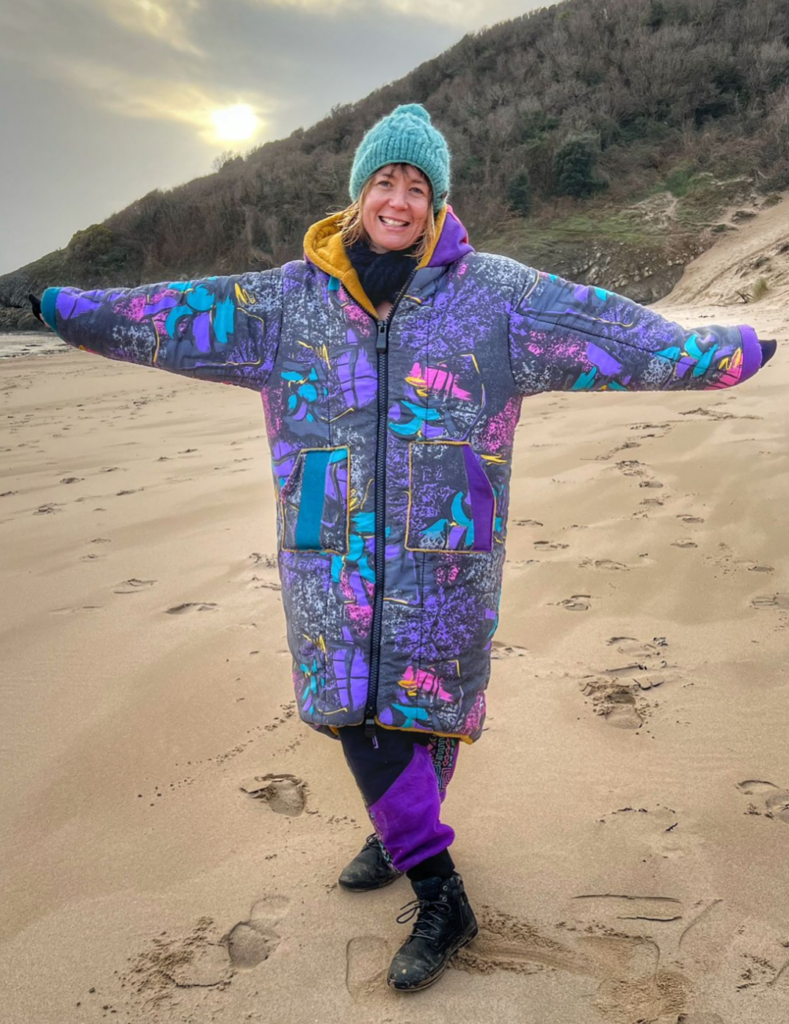 A woman smiling on a beach with a vintage sleeping bag (a fabric patterned with grey, teal, turquoise, purple, pink and yellow).