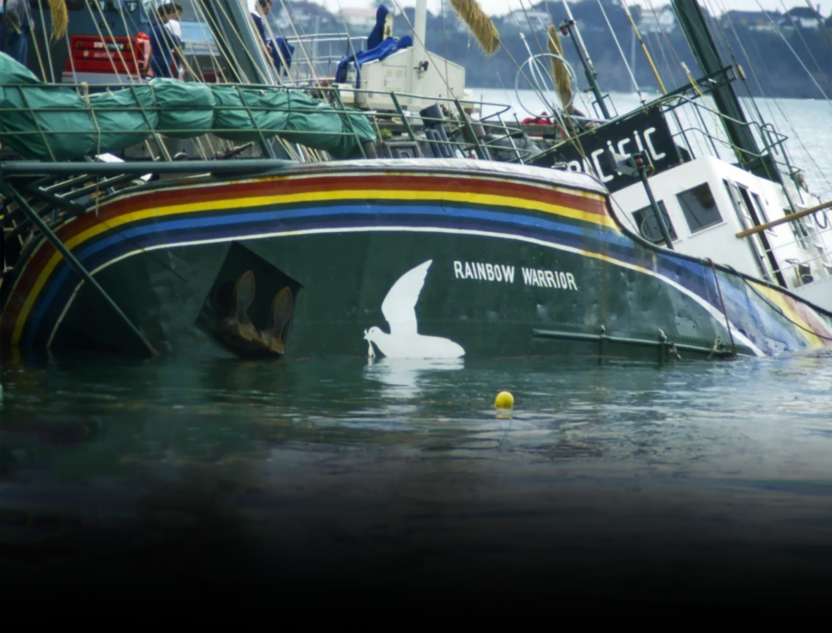 Rainbow Warrior ship sits half-sunk in a harbour. It is tipped to one side and mostly submerged, with the name and rainbow design on the hull still visible.