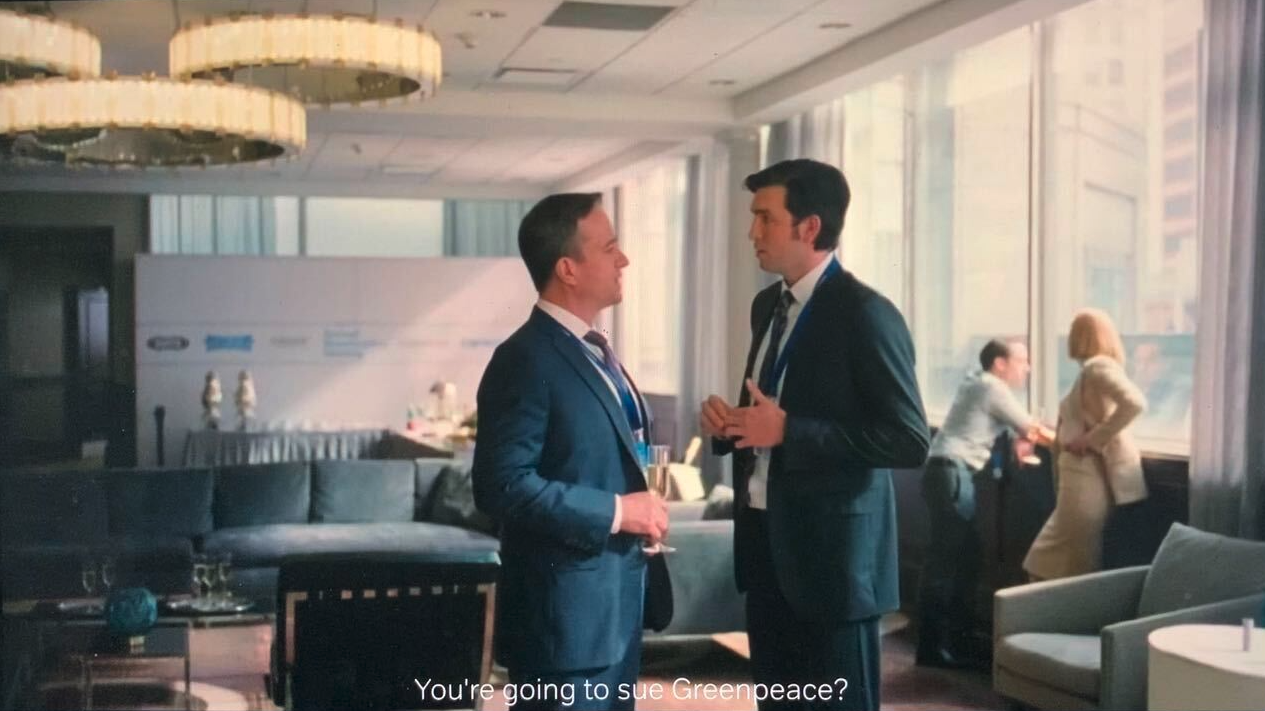 Screenshot from the TV show Succession. Main characters Tom and Greg stand face-to-face in a plush corporate lounge. The subtitle reads 