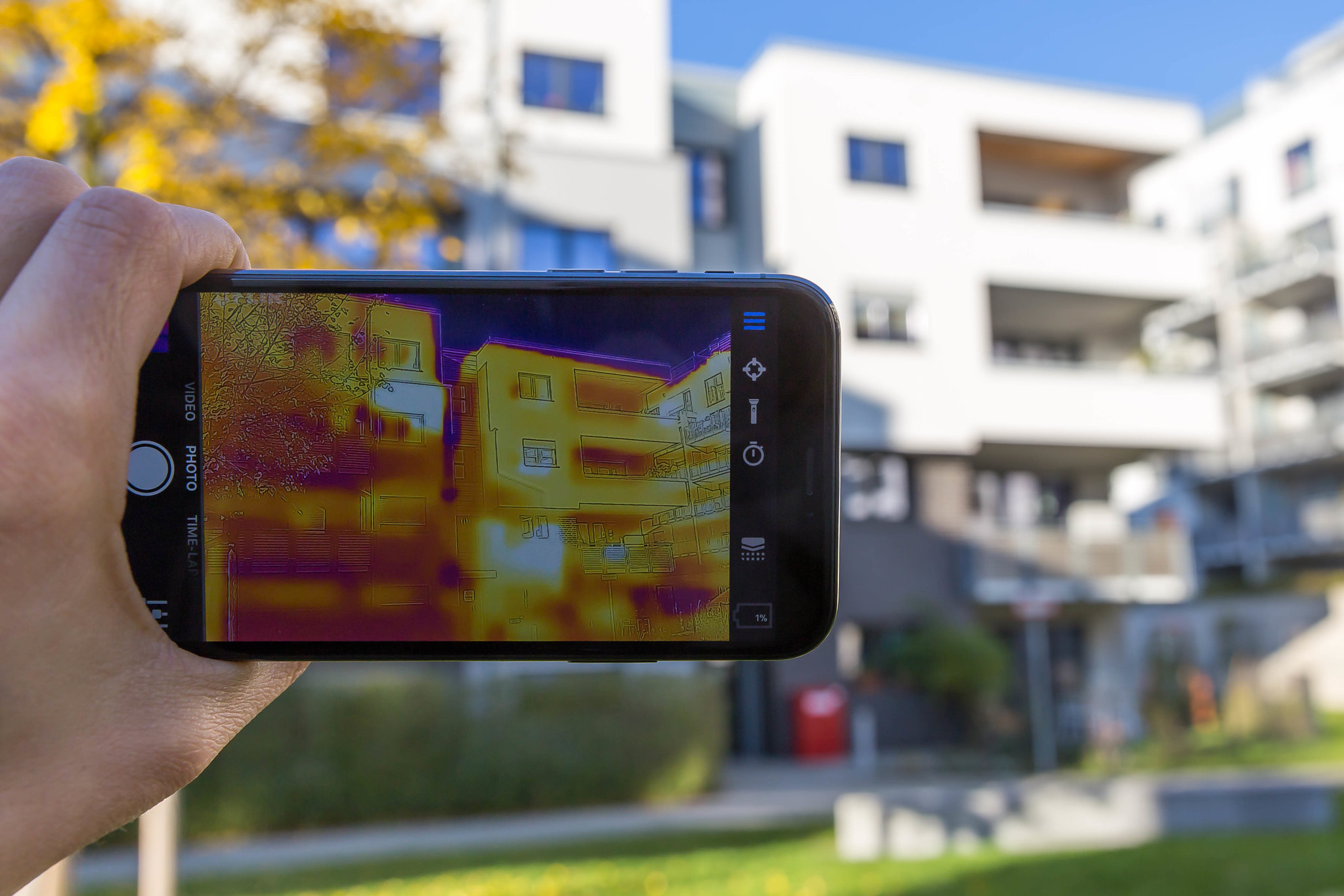 A thermal imaging camera held up to a low-rise apartment building. The camera's screen shows the temperature of different parts of the building's exterior, colour-coded in yellow, orange and purple.