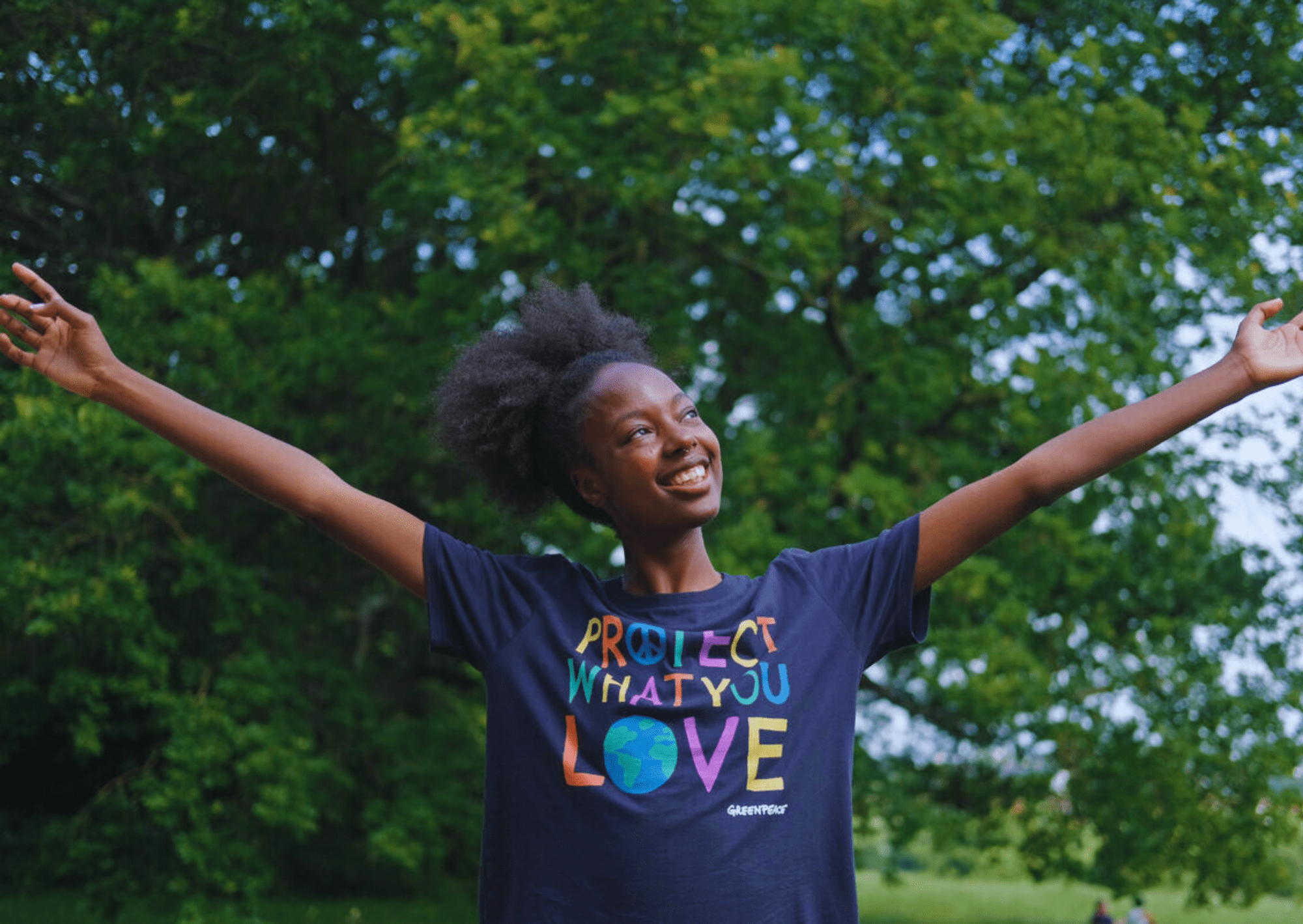 A young smiling Black woman with outstretched arms wearing a dark blue Protect What You Love T-shirt surrounded by trees