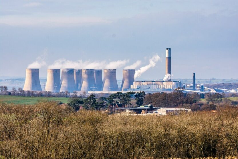 A distant view of several large coal power station chimneys with clouds steaming out of them, and fields in the foreground