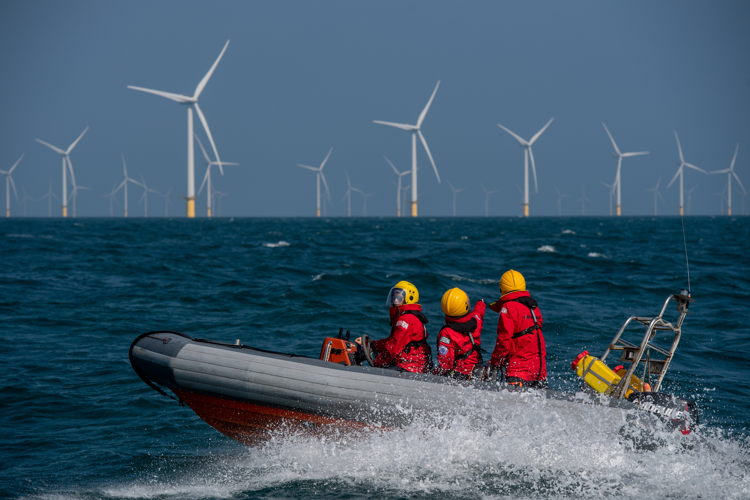 Three Greenpeace crew members in red drysuits and yellow helmets ride a fast-moving inflatable boat across choppy seas, heading towards an offshore wind farm with multiple wind turbines visible on the horizon.