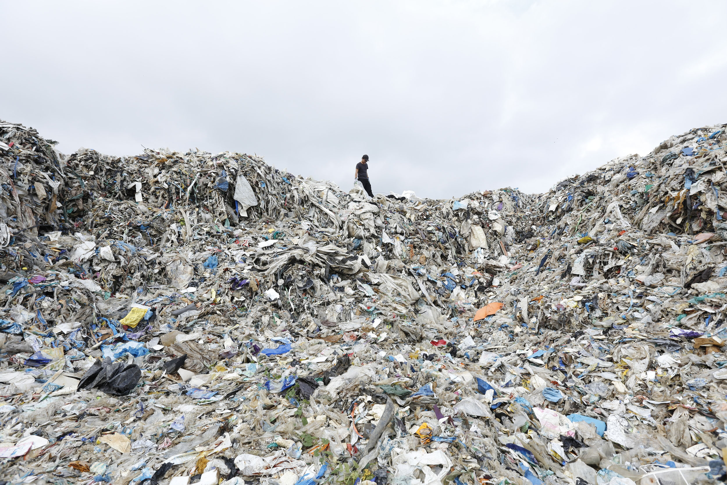A huge heap of waste can be seen in the foreground with a tiny person standing in the background on top.