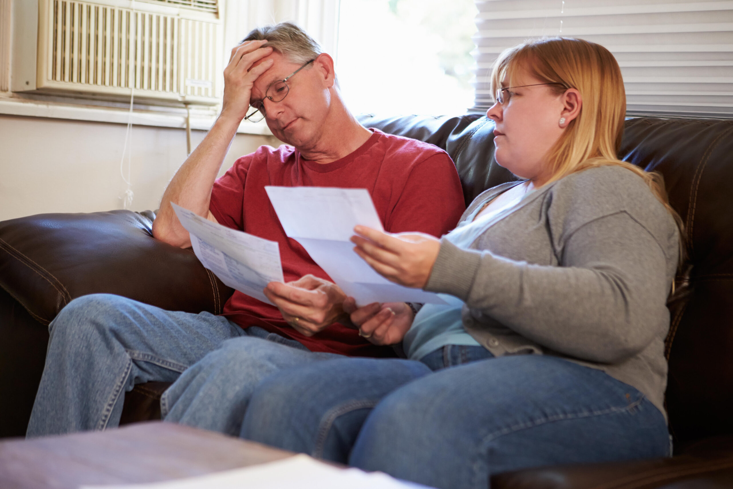 A man and woman sit on a sofa, looking worried as they examine household bills.